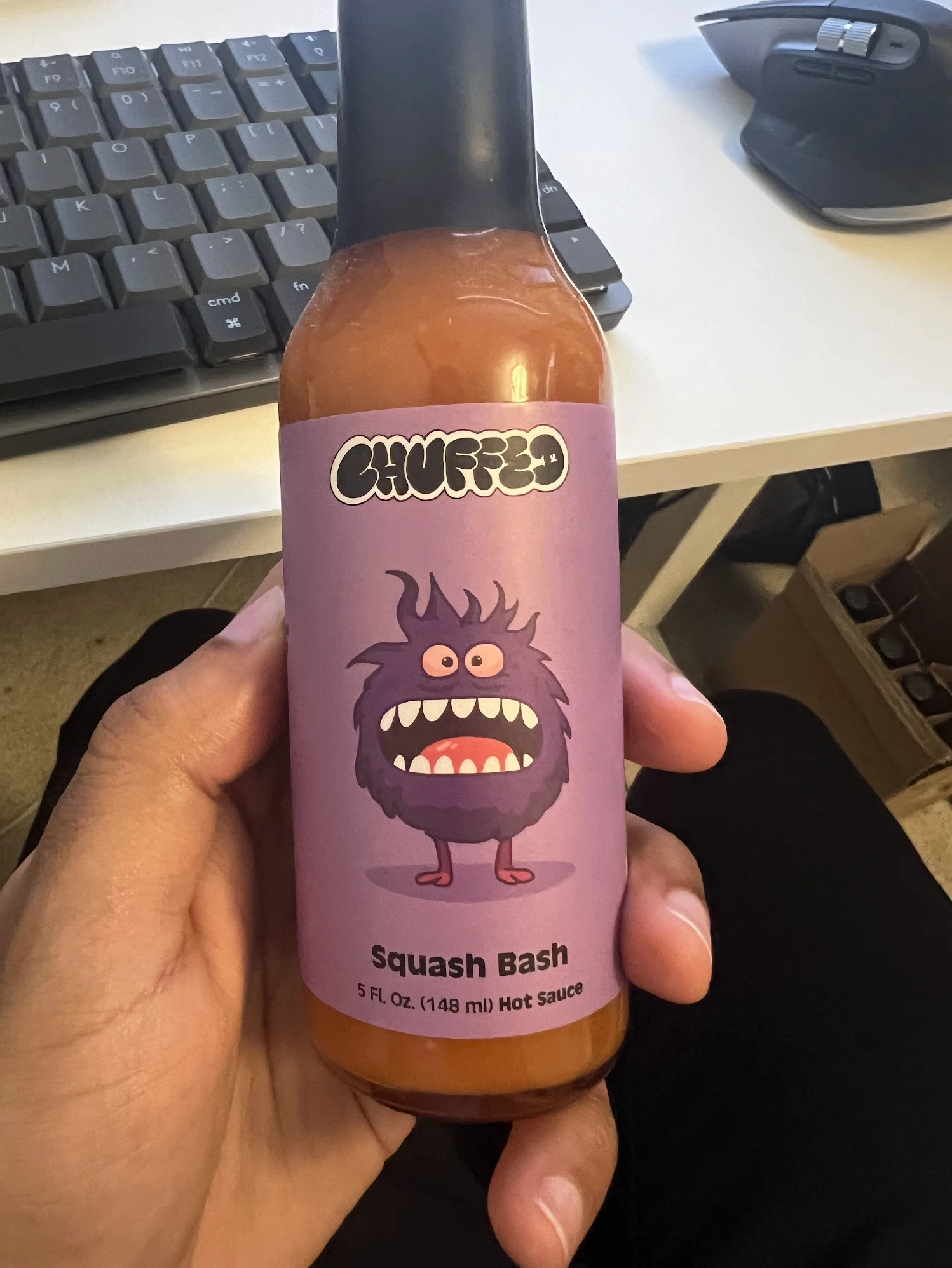 A hand holding a Squash Bash hot sauce sample with a purple character label